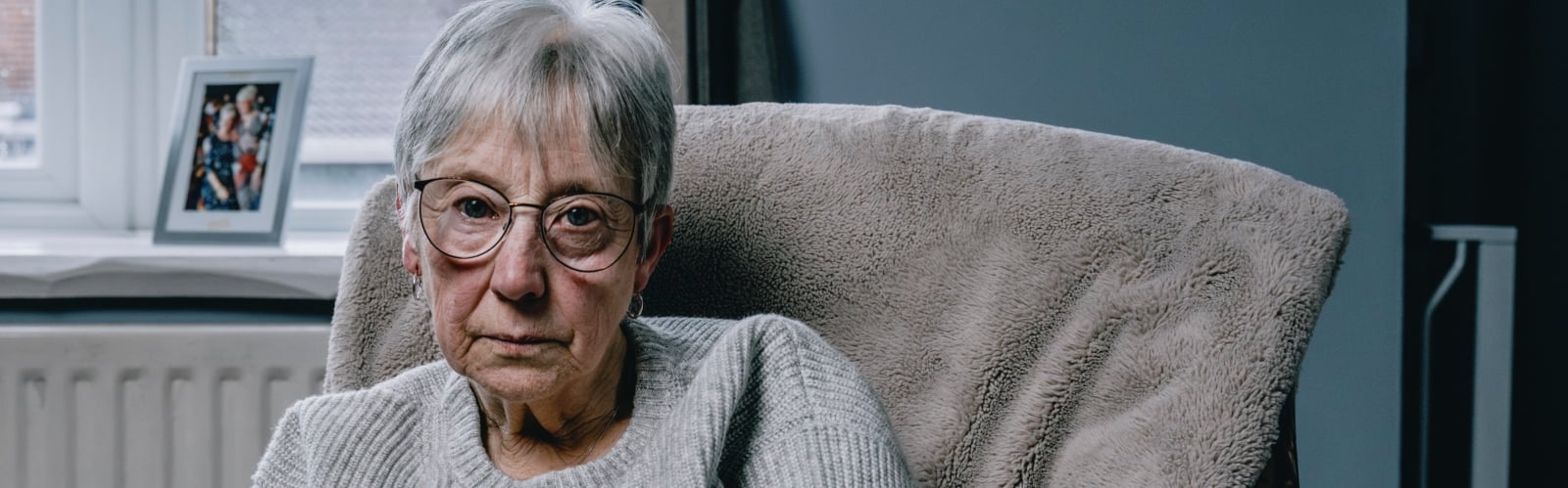A woman with cropped grey hair and glasses looks sadly at the camera